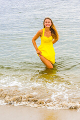 Wearing a bright yellow sundress,  holding up her dress from the bottom, a young pretty woman is wading in water on the beach, smilingly looking forward...