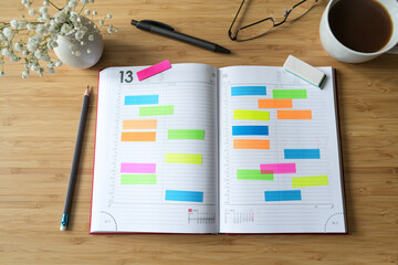 Open diary or appointment calendar with a lot of colorful sticky notes on a wooden desk with coffee pens and glasses, time management concept, copy space, selected focus, flat lay