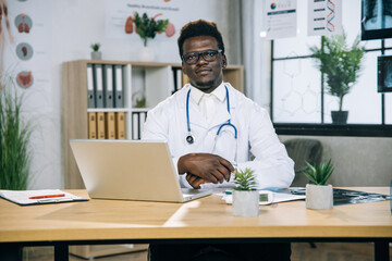 Portrait of african american doctor in eyeglasses and white lab coat sitting at office desk with modern laptop. Medical worker with stethoscope on neck looking at camera. Health care concept.