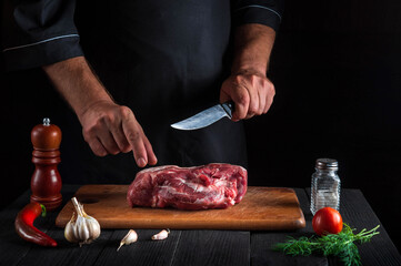 Cook cuts meat with a knife in the kitchen prepares food. Vegetables and spices on the kitchen table in restaurant to prepare a delicious lunch