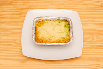Vegetable lasagna with a lot of cheese gratin on top prepared in a metal container fresh from the oven on a light pine wood table