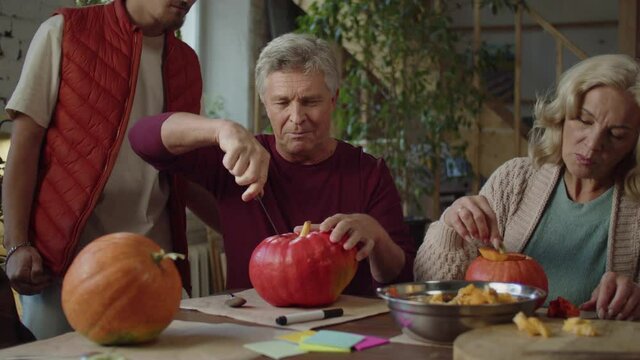 An aged man uses a knife to cut off the top of a pumpkin. The man in the orange yelek helps him