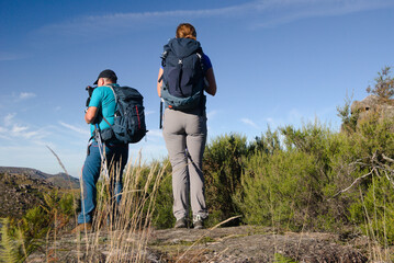 Duas pessoas no topo de uma montanha a ver a vista - uma a fotografar, mochila ás costas -...