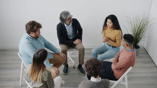 Distressed Asian woman is speaking at group therapy session sharing difficult situations while people sitting in circle are listening and expressing support