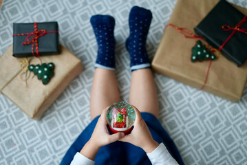 Little girl sitting on the floor with Christmas gifts holding a snowball. View from above