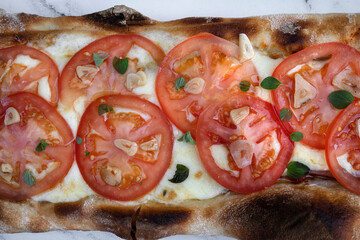 Top view of a traditional Neapolitan pizza with mozzarella cheese, roasted garlic, sliced tomato and fresh oregano leaves, on the white marble table.