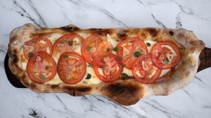 Top view of a traditional Neapolitan pizza with mozzarella cheese, roasted garlic, sliced tomato and fresh oregano leaves, on the white marble table.