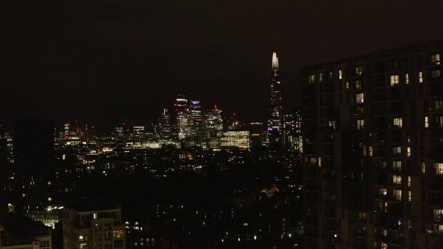 Forwards fly above urban neighbourhood at night. Revealing view of glowing downtown skyscrapers. London, UK