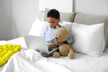 Little African-American boy with laptop and teddy bear watching cartoons in bed