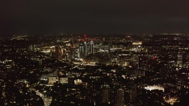 Aerial panoramic view of large city at night. City lights glowing into dark. Fly above urban neighbourhood. London, UK