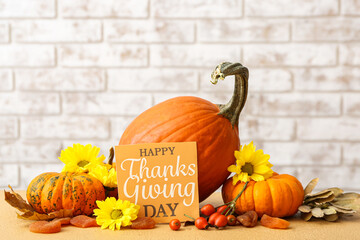 Tasty pumpkins, flowers and text HAPPY THANKSGIVING DAY on table against brick wall