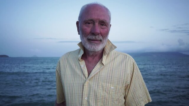 Positive senior man with grey beard and hair wearing yellow checkered shirt stands on sea beach under clear sky in windy evening