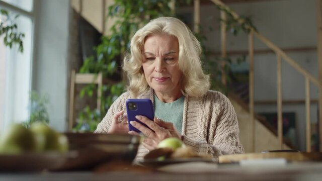 An elderly lady is texting using a smartphone. She sits at the table and smiles