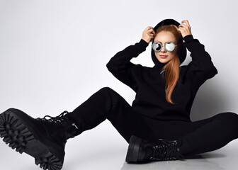 Cool red-haired woman musician in round sunglasses trendy black sports suit and massive brutal shoes sits on floor with legs wide apart, putting on or taking off her cowl