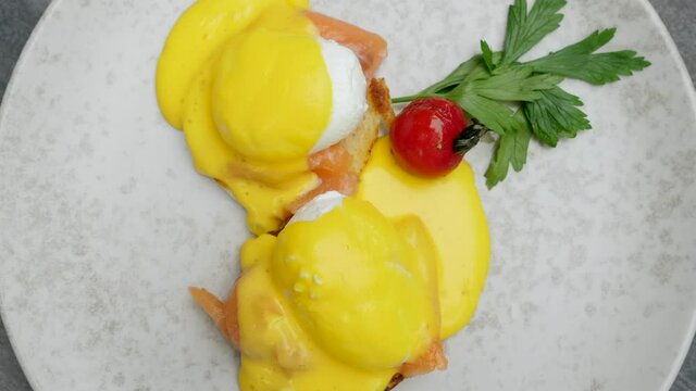 Delicious breakfast with eggs benedict in restaurant. Juicy poached eggs with hollandaise sauce, smoked salmon and cherry tomato in cafe. Plate with prepared food for lunch or dinner. Top view motion