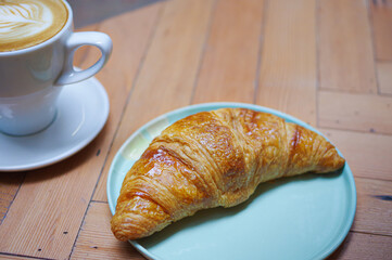 Curved French Croissant in cyan ceramic plate and cup of coffee with rosetta pattern latte art on wooden table top view