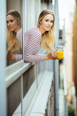 Young woman drinking a glass of natural orange juice, leaning out the window of her home.