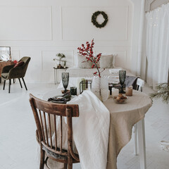 Elegant aesthetic Christmas dinner serving concept. Bright living room interior with chair, table with beige linen table cloth, berries bouquet. Christmas celebration evening
