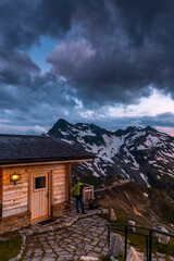 Fototapeta na wymiar Man Watching Sunset from Luxury Cozy Wooden Chalet in Alpine Mountains Peak