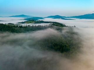A thin morning fog covers the Ukrainian mountains. Green grass on the slopes of the mountains. A curly thin fog spreads over the mountains. Aerial drone view.
