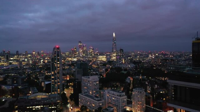 Panoramic view of night city with downtown skyscrapers. Backwards reveal of top of high rise building. London, UK
