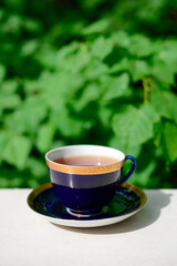 A cup of tea on a white table in the garden, against the background of green leaves of trees. Aromatic and healthy morning or afternoon tea in the open air. Hot tea in a beautiful blue cup with a gold