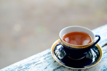 A cup of tea on an old wooden board, an empty blurry background. Black or herbal tea in a beautiful blue cup with a gold rim on the summer veranda or terrace. Afternoon or morning tea