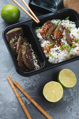 Close-up of smoked tuna slices and white rice in a black plastic delivery container, vertical shot on a grey concrete surface