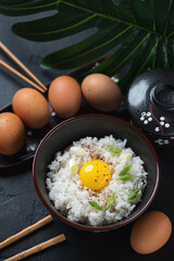 Bowl of japanese warm rice mixed with a raw chicken egg, vertical shot on a black stone background, selective focus