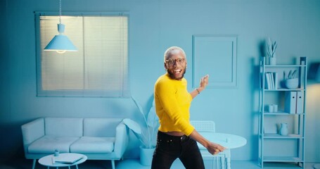 Smiling black man wearing glasses and a yellow shirt dancing at home with blue color background celebrating freedom, excited funky guy listening music enjoying silly movements dance in blue studio