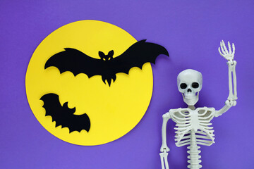Skeleton on purple paper Halloween background and silhouettes of two black paper cut bats flying on...