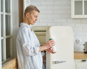 Beautiful senior woman wearing glasses looking at bottle with yogurt, check shelf life and ingridients of product in kitchen near opened refrigerator. Mature people and healthy eating concept