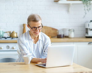 Modern happy mature woman wearing glasses working or studying on laptop while sitting in kitchen at home looking at screen and typing something on keyboard. Senior people and technologies concept