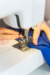Close up shot of tailor or seamstress sewing or stitching blue navy fabric with professional machine while sitting and making new clothing item at her workplace in atelier or workshop, selective focus