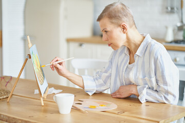 Creative hobby. Side view of concentrated mature woman painting picture at home while sitting at...