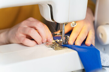 Naklejka premium Close up shot of tailor or seamstress sewing or stitching blue navy fabric with professional machine while sitting and making new clothing item at her workplace in atelier or workshop, selective focus