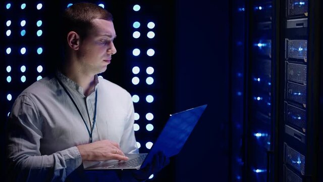 The Concept of Digitalization of Information: IT Specialist Standing In front of Server Racks with Laptop, He Activates Data Center with a Touch Gesture. Network Data