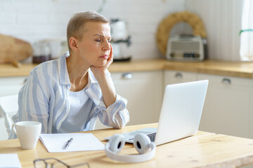 Stressed mature woman in casual wear sitting in modern kitchen, holding head in hands and looking on laptop screen while working or studying online from home. Senior people and technologies concept