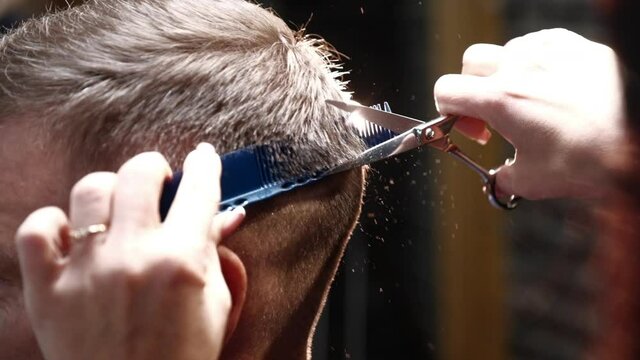 Professional unisex hairdresser with neat manicure cuts man short dark hair on back using steel scissors and comb in salon closeup