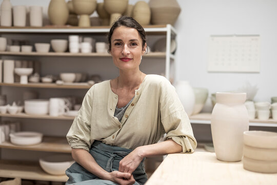 Portrait of beautiful happy craft woman wearing apron and smiling while sitting in her art studio or craft pottery shop