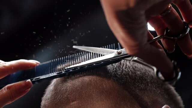 Professional unisex hairdresser cuts man short dark hair on hindhead using scissors and comb in salon extreme closeup slow motion