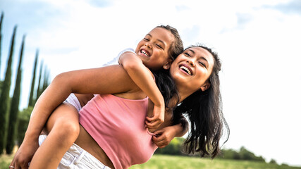 Asian mother and daughter having fun outside - Portrait of happy woman giving a piggyback ride to...