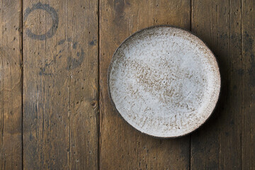 Clean empty ceramic textured plate on dark brown wooden table background. Horizontal, copy space, top view. Concept for menu with utensil. Mockup of round earthware cutlery