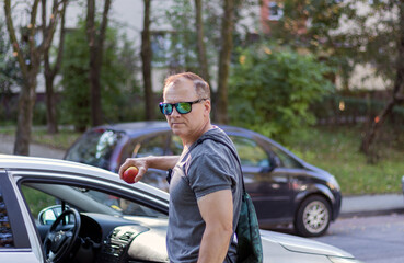 mature man, handsome courageous, 50 years old, dressed in a T-shirt, stands near the car and holds an apple, a man's portrait on a city background