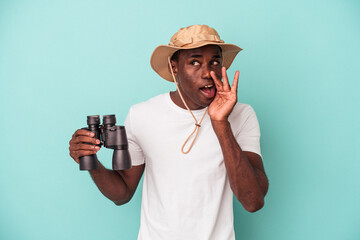 Young African American man holding binoculars isolated on blue background is saying a secret hot braking news and looking aside