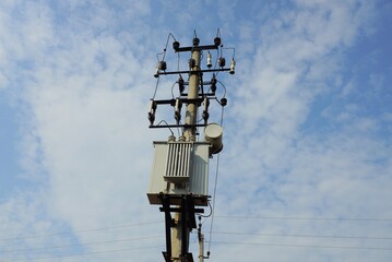gray metal transformer on a pole with electric wires against a blue sky and white clouds
