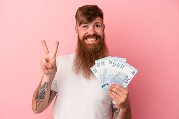 Young caucasian ginger man with long beard holding banknotes isolated on pink background showing...