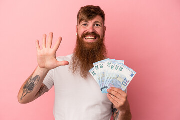 Young caucasian ginger man with long beard holding banknotes isolated on pink background smiling...