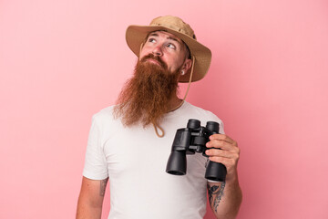 Young caucasian ginger man with long beard holding binoculars isolated on pink background dreaming of achieving goals and purposes