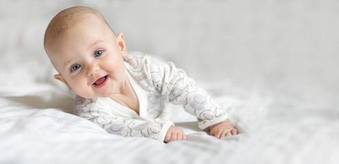 Happy little toddler baby girl smiling lying on the bed on a white sheet at home looking at the camera. Copy space for text, mock up, banner - obrazy, fototapety, plakaty